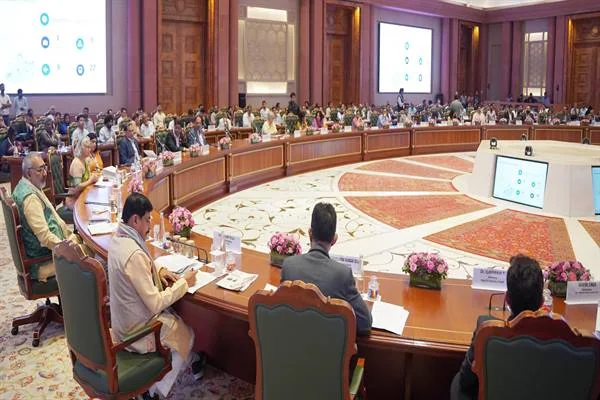 बड़े गोल सम्मेलन मेज पर बैठे अधिकारी, स्क्रीन और फूल, मुख्यमंत्री एक कुर्सी पर